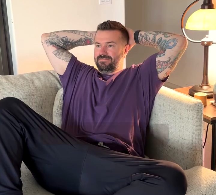 A man stretched out on his couch beside a lamp, hands behind his head