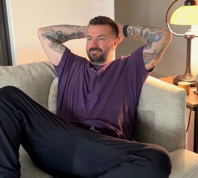A man stretched out on his couch beside a lamp, hands behind his head