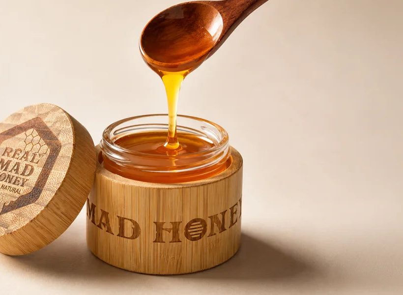 A wooden spoon of dark honey dripping into an open bamboo jar of Real Mad Honey